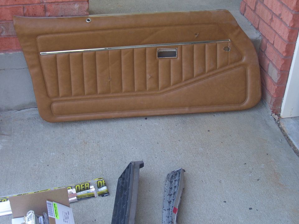 Tan vinyl door panel with horizontal stitching and chrome trim strip, lying on concrete. Two other car parts are visible below