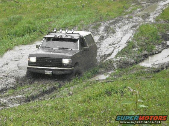 Muddy dark Ford Bronco with white roof and roof light bar driving through a rutted mud pit