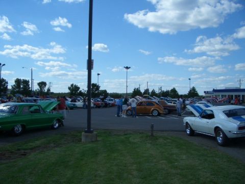 1970 Ford Mustang - A gathering of vehicles in a parking lot on a partly cloudy day.