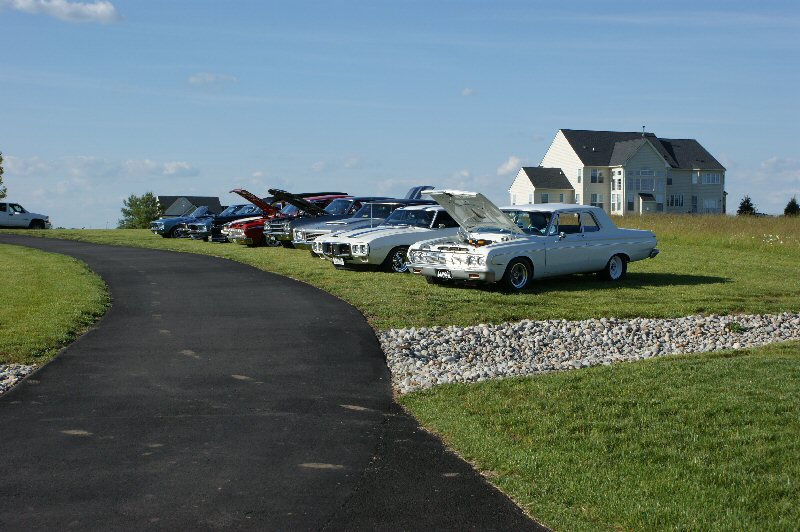 several old cars lined up on grass with hoods open, house in background