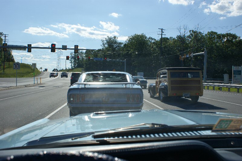 Rear view of a 1970 Ford Mustang driving on a sunny day, with a wood-paneled station wagon in the right lane