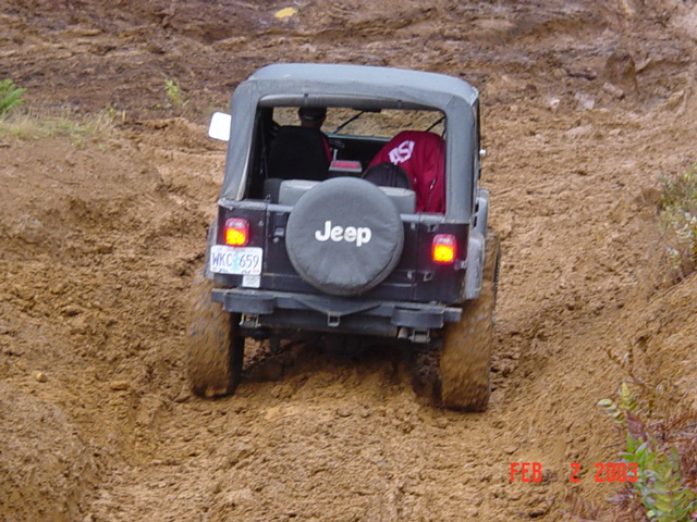 Jeep Wrangler driving on a dirt trail, rear view. It has a soft top and a spare tire on the back