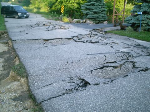 2001 Dodge 2500 - A black van is parked on a road that has sustained significant damage, with large cracks and sections of pavement collapsed.