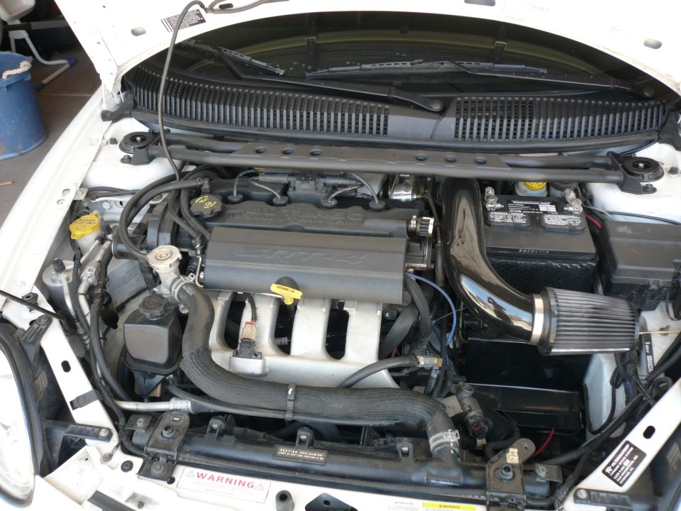 White car engine bay with SRT-4 branding on the engine cover, aftermarket air intake, and battery