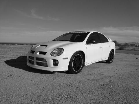 1996 Ford Probe - White Dodge Neon SRT-4 with a hood scoop and black wheels is parked on a dirt lot under a cloudy sky.