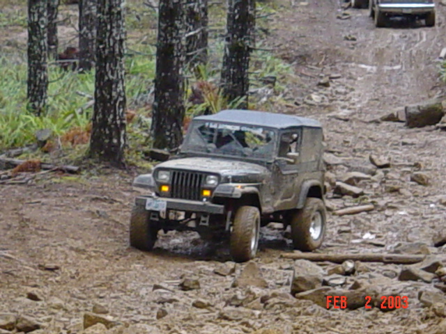 Muddy Jeep Wrangler with cracked windshield on a rocky, wooded trail