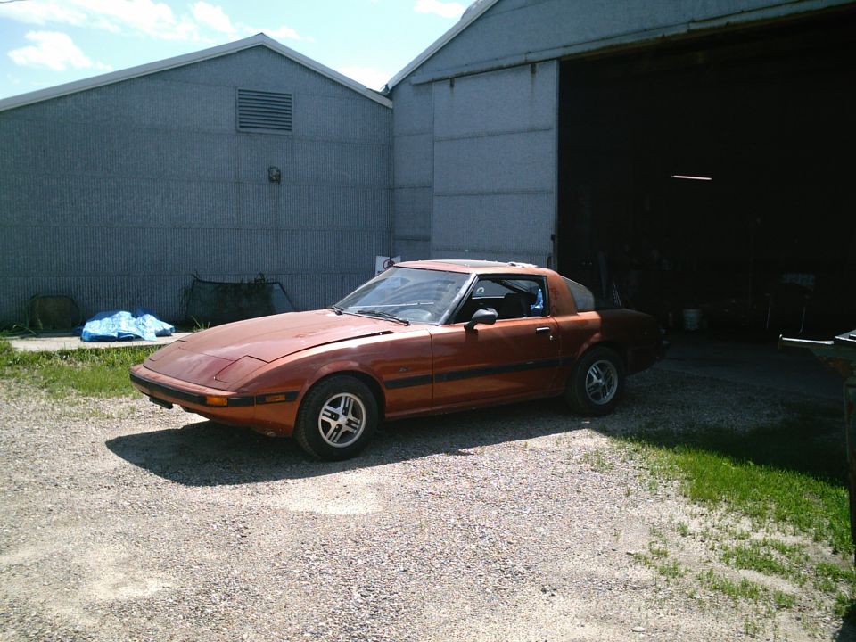 Copper Mazda RX-7 coupe parked on gravel by an open industrial garage door.