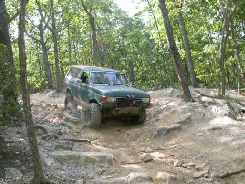 1990 Ford Bronco - A green 1990 Ford Bronco navigates a challenging rocky path through a wooded area.