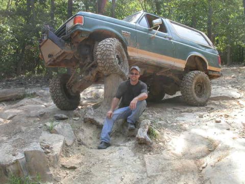 1990 Ford Bronco - A man sits in front of a lifted green 1990 Ford Bronco on a rocky, wooded trail.