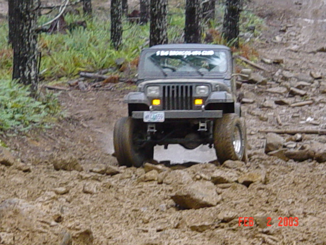 1995 Jeep Wrangler driving through mud and rocks on a wooded trail