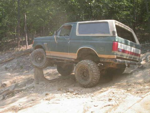1990 Ford Bronco - A green 1990 Ford Bronco navigates a challenging off-road course with its front wheel elevated on a rock.