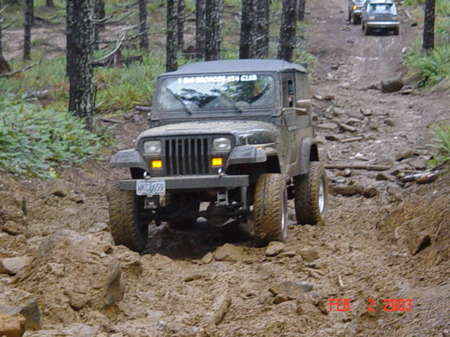 Jeep Wrangler driving on a rocky, muddy trail with trees on the side. Another vehicle is visible further up the trail