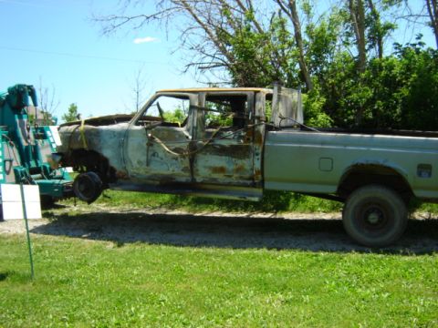 1992 Ford Bronco - A heavily damaged pickup truck is lifted by a tow truck, showing extensive rust and missing parts.