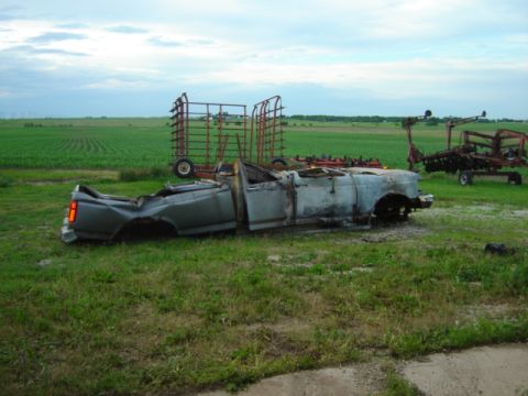 1992 Ford Bronco - This heavily damaged pickup truck sits in a field, its body showing significant fire or impact damage.