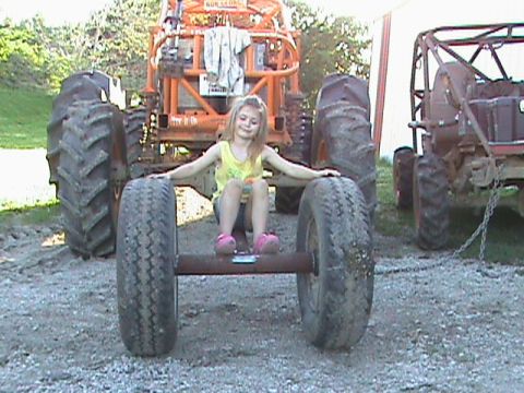 1973 Ford Bronco - Girl sits on an axle between the massive tires of a tractor, with another vehicle visible to the right.