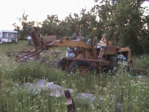 1973 Ford Bronco - A yellow front loader sits in a field, partially obscured by tall grass and weeds, with a person standing on its platform.