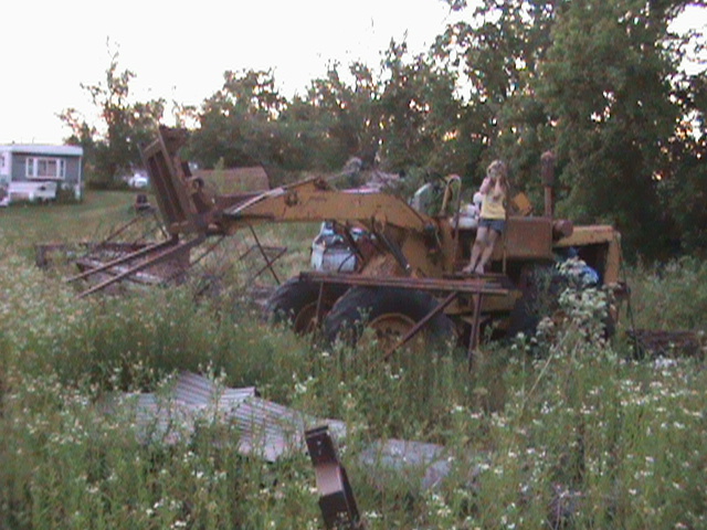 Yellow front loader with a person standing on it, overgrown with weeds
