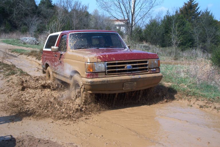 1989 Ford Bronco driving through a muddy trail, kicking up water and mud