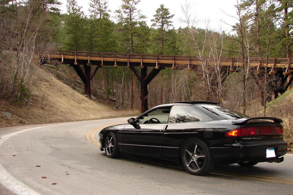 Black Ford Probe coupe rear 3/4 view parked on a curving road near a wooden trestle bridge.