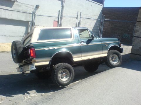 1985 Ford Bronco - A green and tan Ford Bronco is parked at an angle on a curb, with a spare tire mounted on the rear.