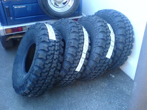 1985 Ford Bronco - Four new off-road tires are lined up next to a blue vehicle.