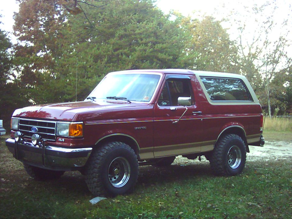 Red and tan Ford Bronco SUV with brush guard and large off-road tires parked on grass, front 3/4 view