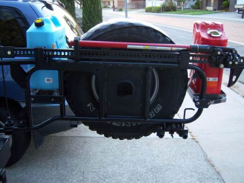 1991 Ford Bronco - The rear of a blue SUV is shown with a spare tire mounted on a black swing-away carrier. A red jerry can and a blue water jug are attached…