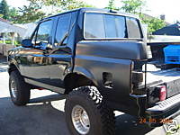 1991 Ford Bronco - The rear three-quarter view of a modified black Ford Bronco is shown. The vehicle has large off-road tires, a lifted suspension, and a cust…