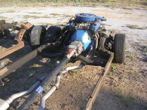 1975 Ford Ranchero - A disassembled vehicle chassis with a blue engine and exhaust system is shown outdoors on a gravelly surface. The front wheel and part of t…