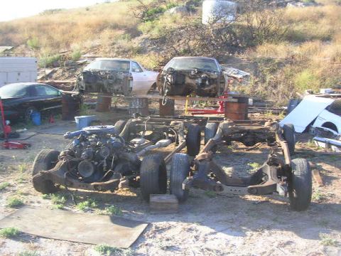 1975 Ford Ranchero - The chassis and engine of a disassembled vehicle are in the foreground, with two car bodies elevated on barrels in the background. A black…