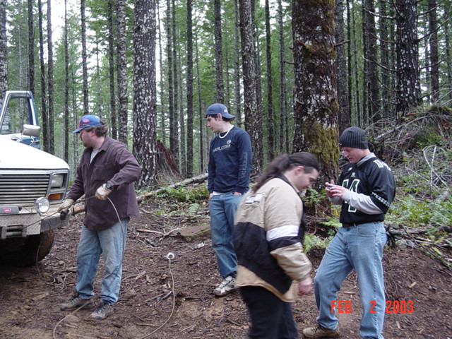 Man in brown shirt attaches a cable to the front of a white vehicle in a wooded area; three other people stand nearby
