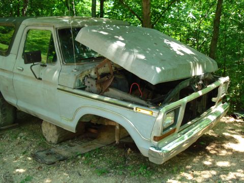 1989 Ford Bronco - A light-colored Ford Bronco with its hood open is parked outdoors in a wooded area. The vehicle appears to be in a state of disrepair, supp…