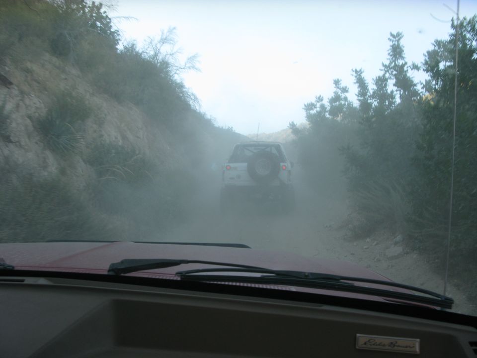 View from inside a vehicle looking at a white Ford Bronco driving on a dusty dirt road.