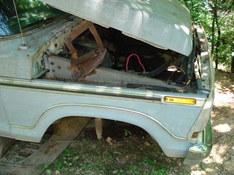1989 Ford Bronco - The front fender and hood of a light blue vehicle are shown, with the hood propped open revealing the engine bay and rusty hinges. The vehi…