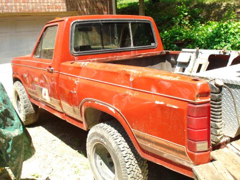 1989 Ford Bronco - The rear passenger side of a faded red Ford pickup truck is shown parked in front of a brick building. The truck bed has a diamond plate to…
