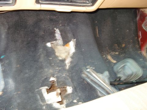 1989 Ford Bronco - The interior floor of a vehicle shows significant wear and tear, with large holes in the black carpet revealing the underlying structure. A…