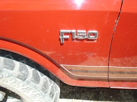1989 Ford Bronco - Close-up of the "F150" badge on the side of a red Ford truck, showing a portion of the tire and a pinstripe detail along the lower body.