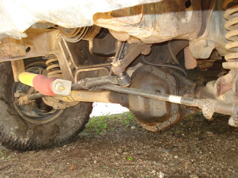 1989 Ford Bronco - The underside of a vehicle shows the front axle, steering components, and a portion of the tire. The steering stabilizer shock is visible w…
