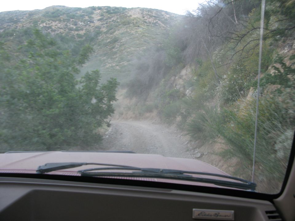 View from inside a vehicle driving on a dusty dirt road in a mountainous area.