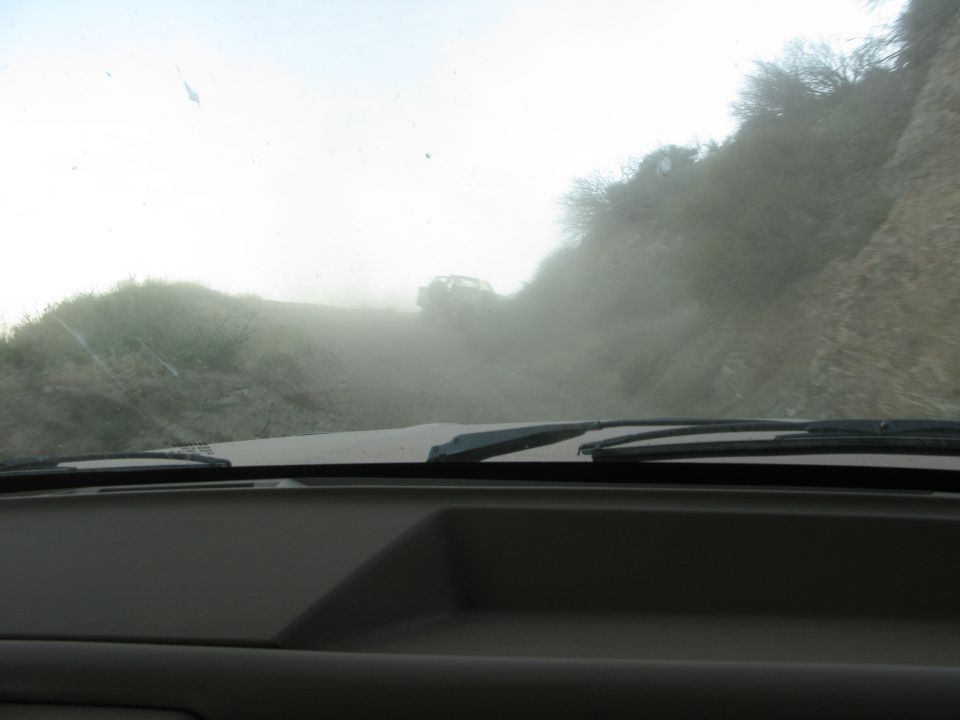 View from inside a vehicle looking out at a dusty dirt road with another vehicle ahead.