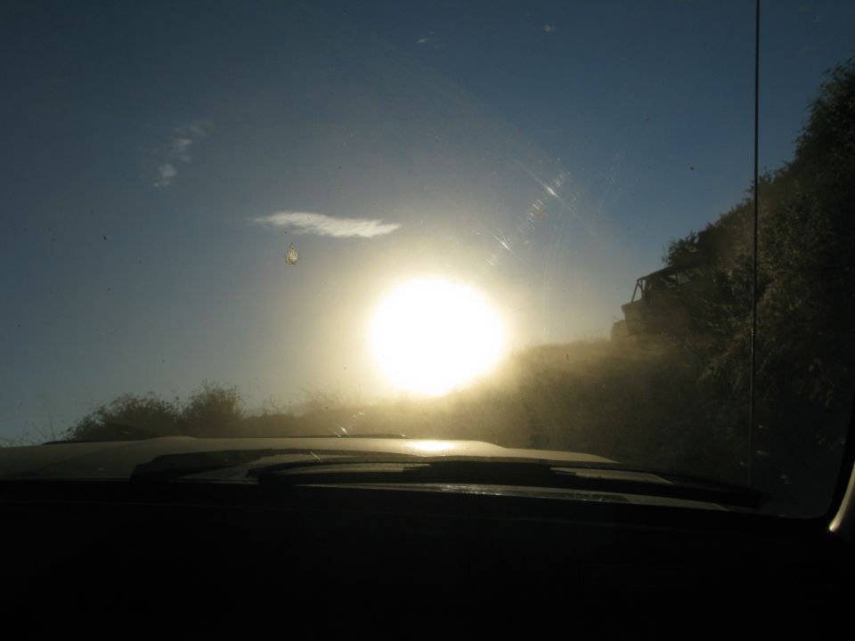 View through a dusty windshield at sunset, with a vehicle on a dirt road ahead.