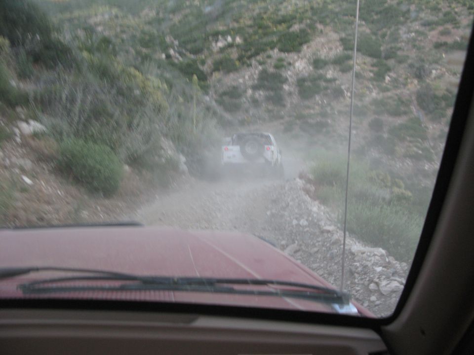 White SUV driving on a dusty mountain road, viewed from inside another vehicle.