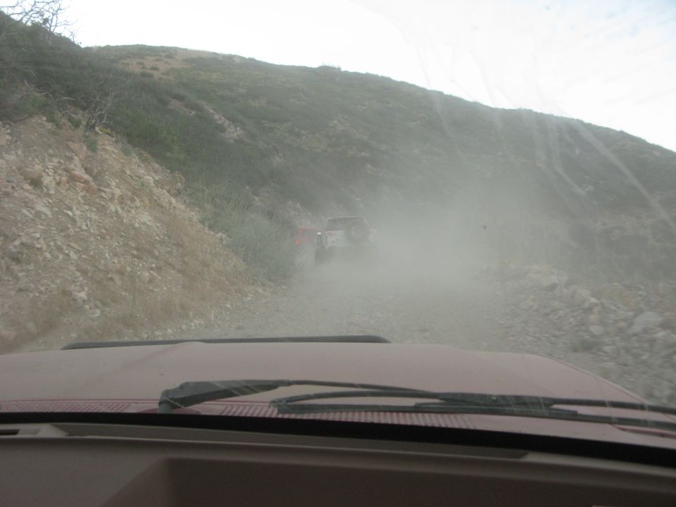 View from inside a red vehicle on a dusty road with two off-road vehicles ahead.