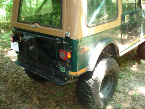 1989 Ford Bronco - The rear passenger side of a green and tan Jeep CJ is shown parked on a dirt path with fallen leaves. The vehicle has a spare tire mounted…