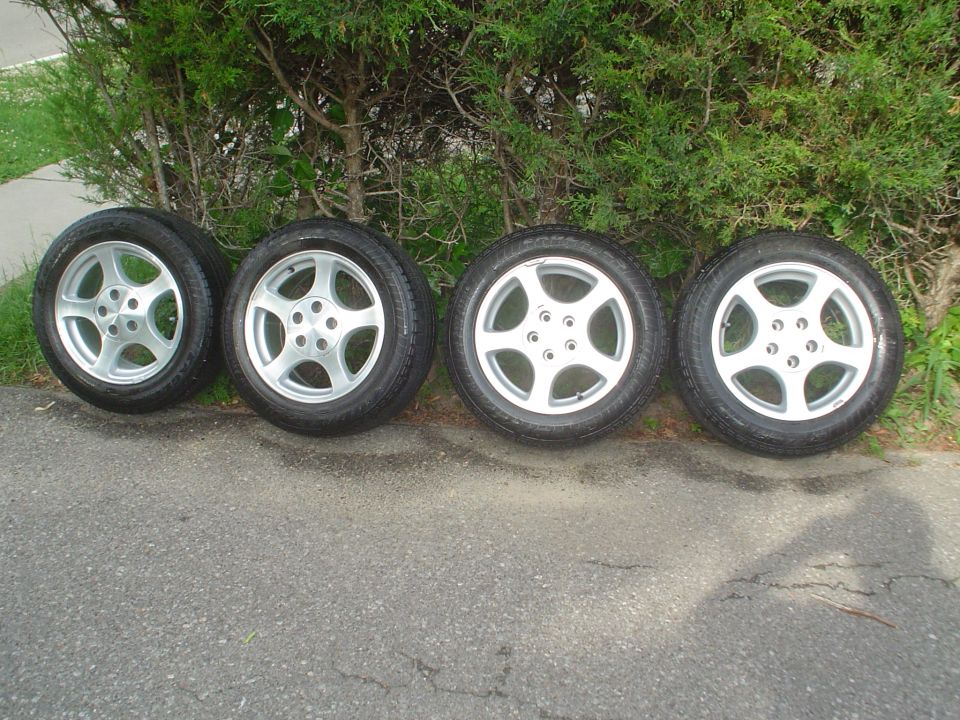 Four silver alloy wheels with tires displayed next to a green hedge.