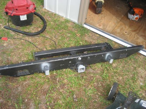 1981 Ford Bronco - A black steel rear bumper with an integrated square receiver hitch tube and mesh light cutouts lies face-up on the grass outside a metal sh…