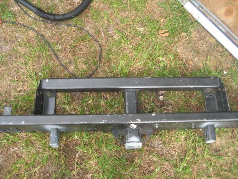 1981 Ford Bronco - A black steel tube bumper lying face-up in the grass, built as a two-rail ladder frame with bolt-through mounting plates at each end and a…