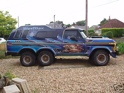1991 Ford Bronco - A custom blue six-wheeled Ford Bronco with elaborate graphics and the name "BlueMax" on the roof. The vehicle is parked on a gravel surface…