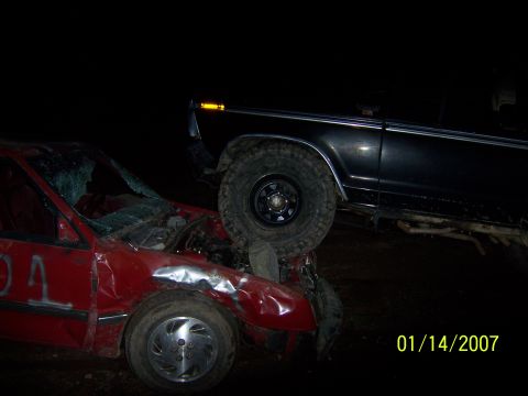 1989 Ford Bronco - A damaged red car is partially crushed by a black SUV at night. The SUV's front tire rests on the red car's hood and windshield.