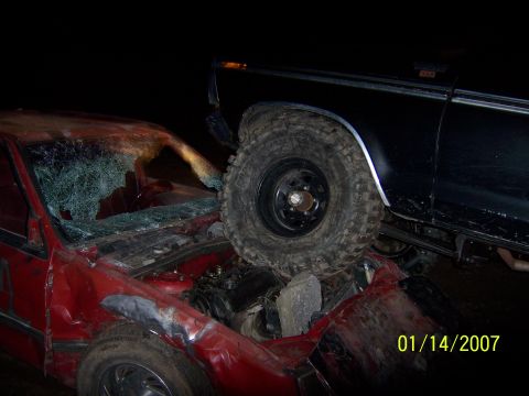 1989 Ford Bronco - A heavily damaged red car is crushed underneath the large tire of a black vehicle, likely a truck or SUV, at night. The red car's windshiel…